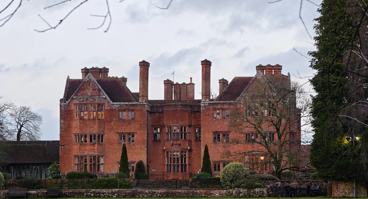 A large brick manor house seen across a green field. The skies are grey and serious looking.